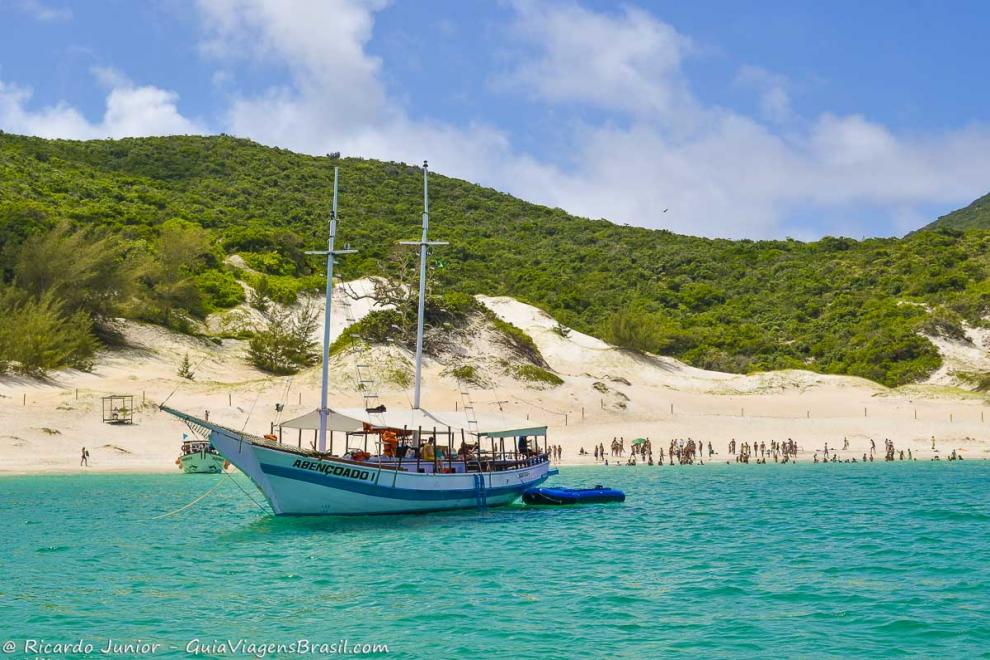 Imagem de turistas na praia e barco de passeio no mar em Arraial do Cabo.